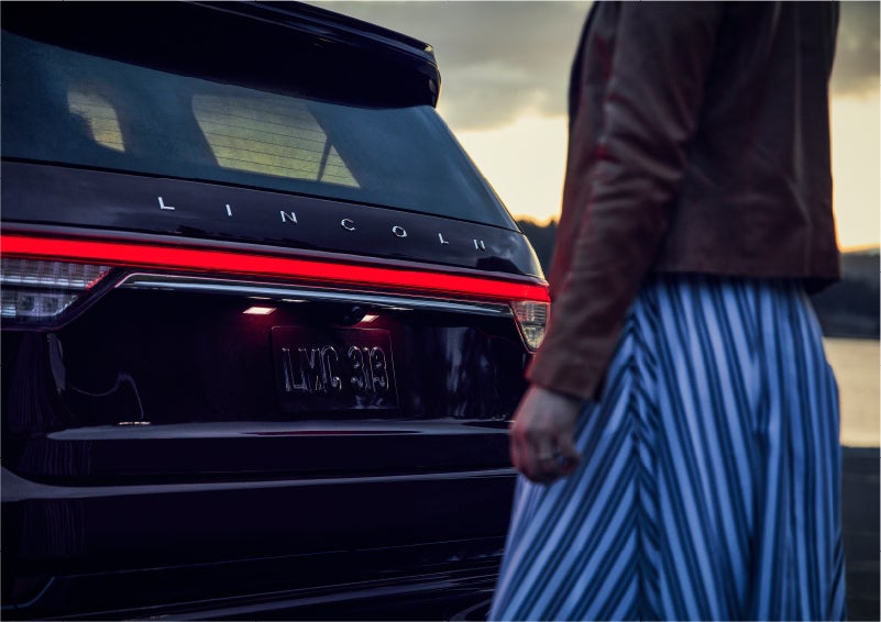 A person is shown near the rear of a 2023 Lincoln Aviator® SUV as the Lincoln Embrace illuminates the rear lights | Empire Lincoln of Huntington in Huntington NY