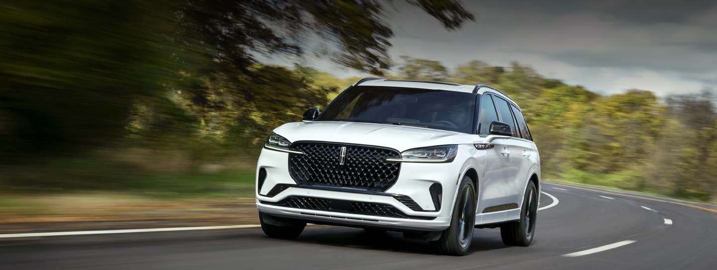 A white 2025 Lincoln Aviator® SUV is being driven down a road lined with trees. | Empire Lincoln of Huntington in Huntington NY